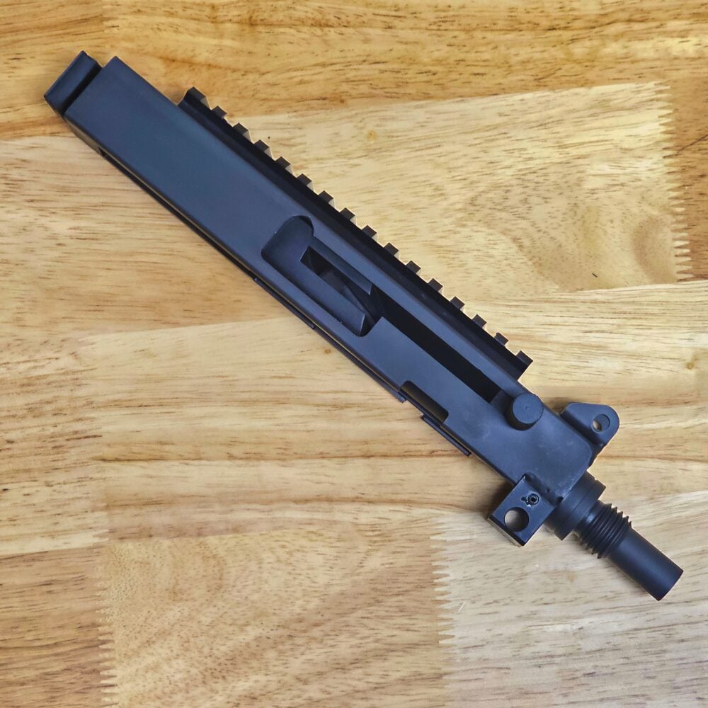 Black rifle upper receiver with Picatinny rail, lying diagonally on a wooden table.