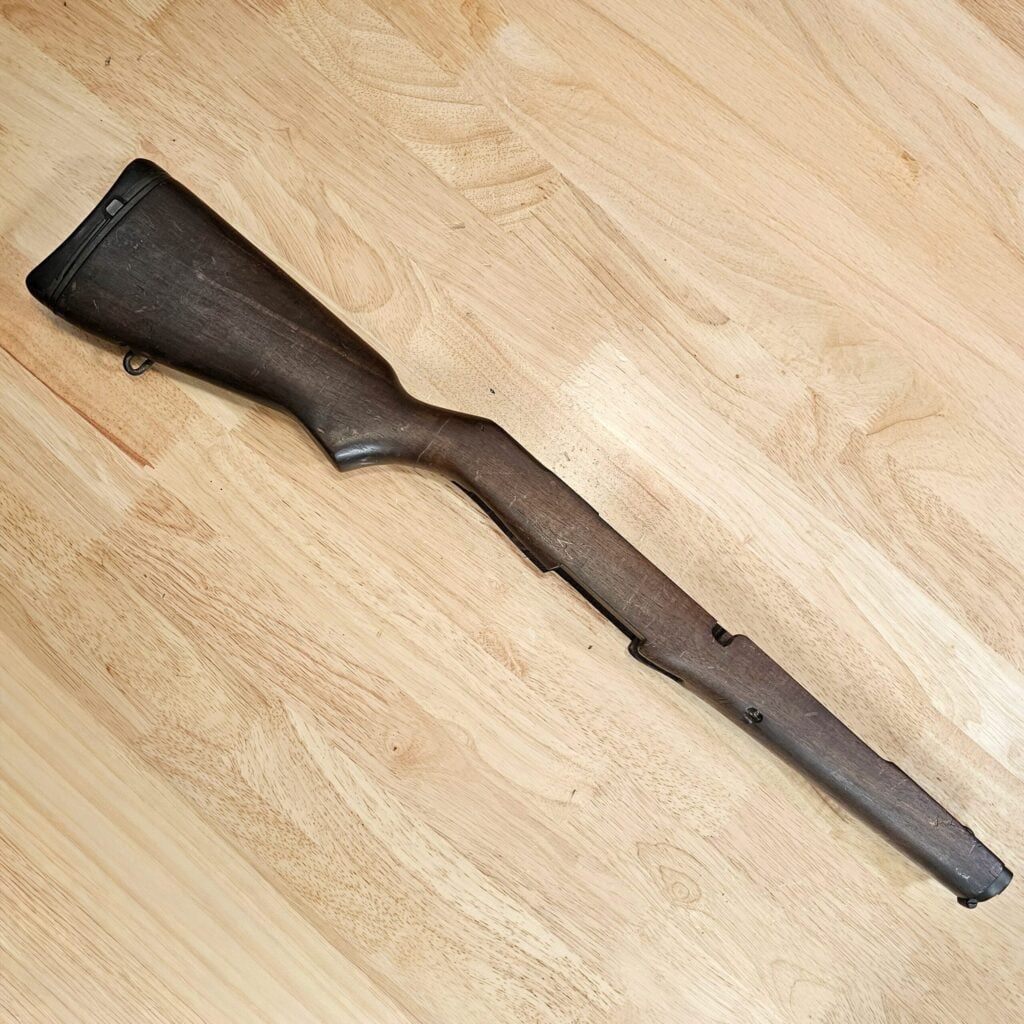 Close-up of a dark wooden rifle stock lying diagonally on a light wooden floor.