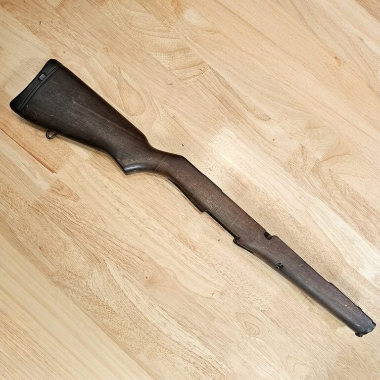 Close-up of a dark wooden rifle stock lying diagonally on a light wooden floor.