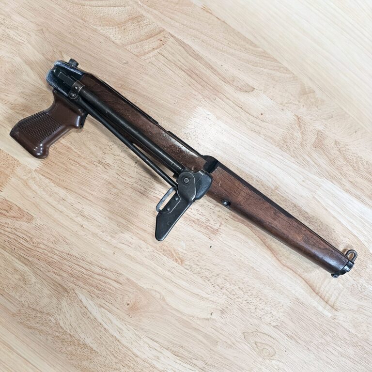 Partially visible rifle with wooden stock resting on a light wooden floor on a diagonal.