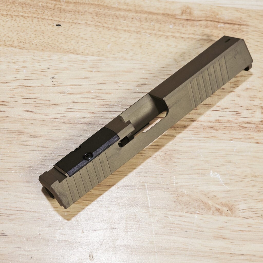 Close-up of a gray pistol slide with serrated sides lying on a wooden workbench surface.
