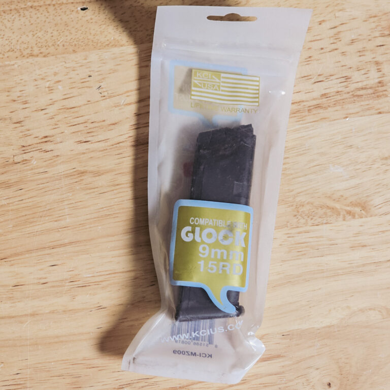 Packaging for a Glock 9mm accessory; clear blister pack with a black part inside and a label reading 'KCI USA Lifetime Warranty' on a wooden surface.