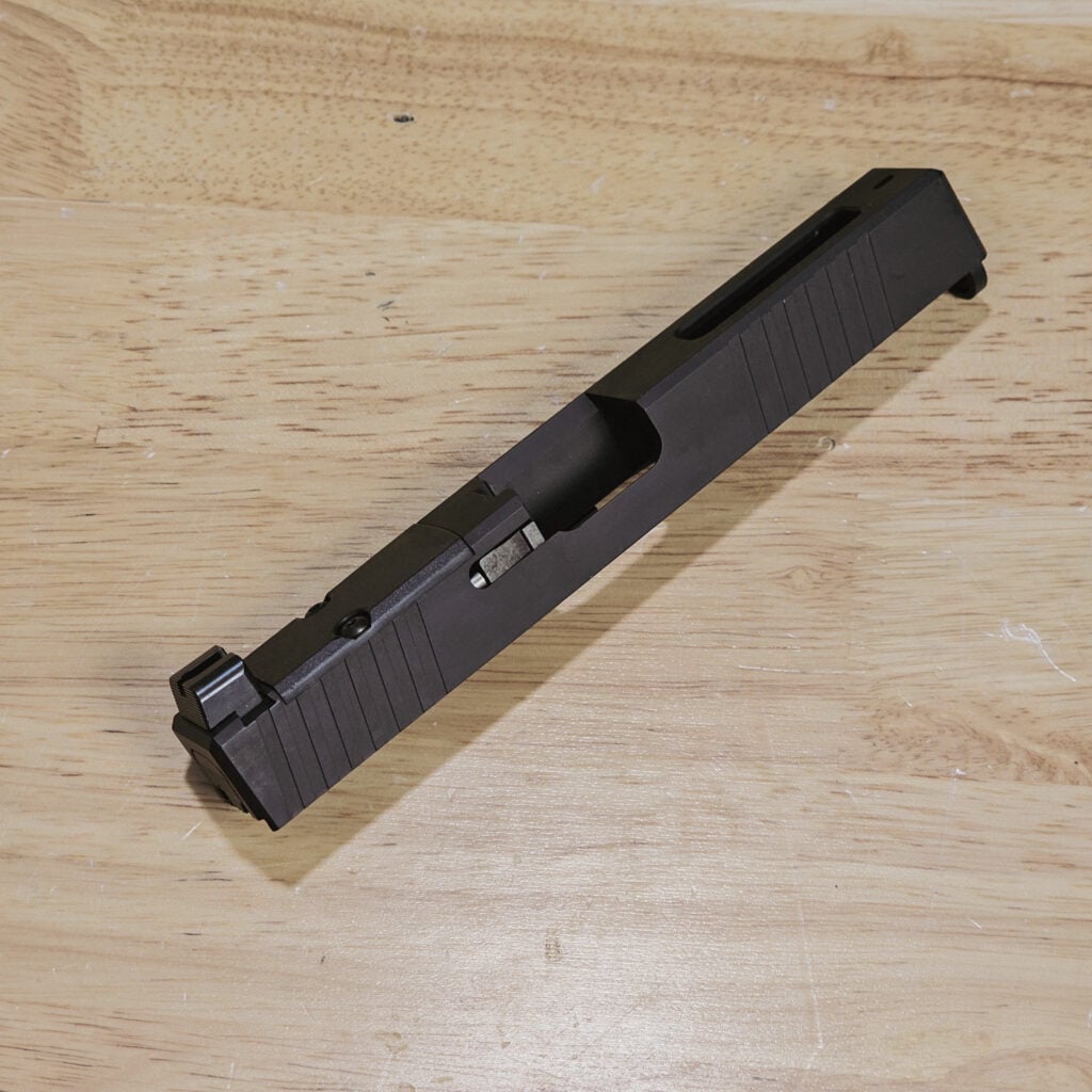 Close-up of a black pistol slide resting on a wooden surface, showing textured grooves along its side.