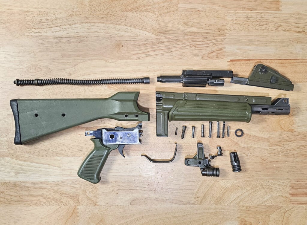 Disassembled rifle parts laid out on a wooden table, including a green stock, barrel assembly, and receiver pieces.