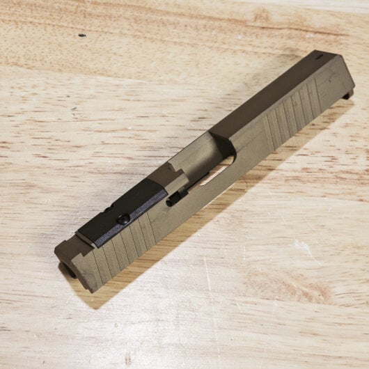 Close-up of a gray pistol slide with serrated sides lying on a wooden workbench surface.